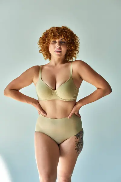 Redhead plus size woman in beige underwear with hands on hips on grey backdrop, body confidence — Stock Photo
