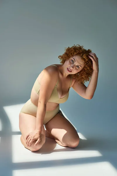 Séduisante femme plus taille en sous-vêtements beige touchant cheveux roux ondulés au soleil sur fond gris — Photo de stock
