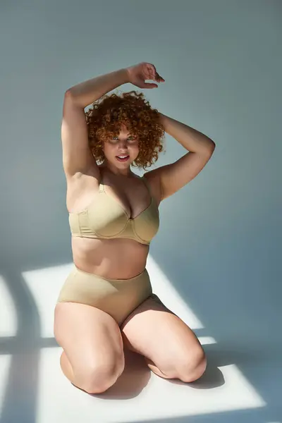 Sensual and redhead plus size woman in beige lingerie looking at camera in sunlight on grey — Stock Photo