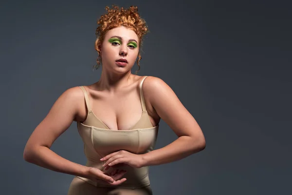 Affascinante modello plus size con capelli rossi ondulati in posa in biancheria intima beige su sfondo grigio scuro — Foto stock