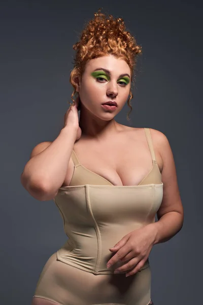 Affascinante rossa e donna curvy in biancheria intima taupe toccare il collo e in posa su grigio scuro — Foto stock