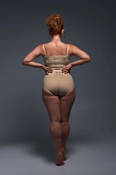 Rückseite der kurvenreichen rothaarigen Frau in beiger Dessous mit Händen an der Taille vor dunkelgrauem Hintergrund — Stockfoto