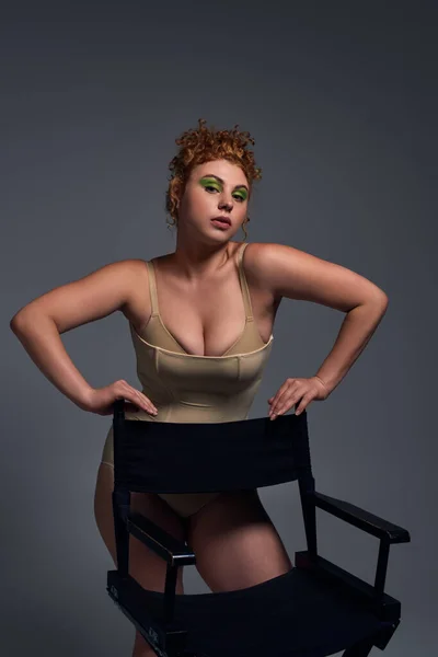Redhead plus size model in taupe underwear posing with chair on dark grey backdrop, body confidence — Stock Photo