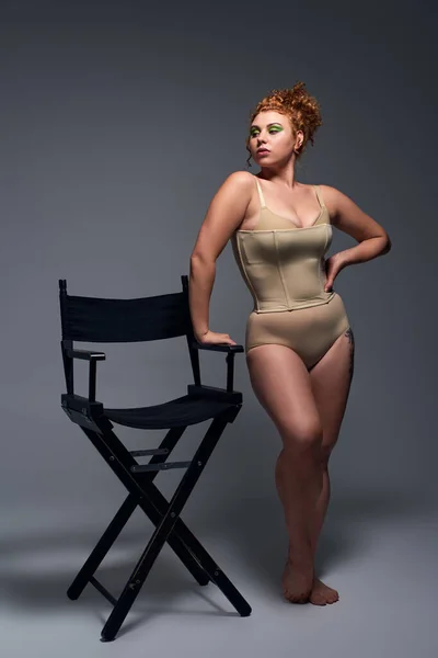 Rossa plus size modello in biancheria intima beige posa vicino alla sedia e guardando lontano su sfondo grigio scuro — Foto stock