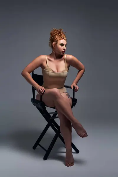 Full length of young redhead woman with curvy body sitting on chair in beige lingerie on dark grey — Stock Photo