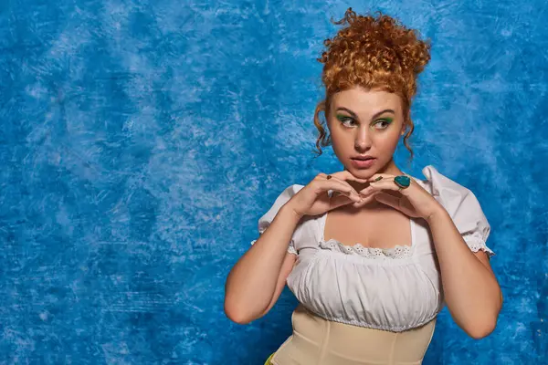 Elegant redhead plus size model holding hands near chin and looking away on blue textured backdrop — Stock Photo