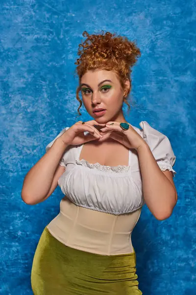 Stylish redhead plus size model with hands near chin looking at camera on blue textured backdrop — Stock Photo