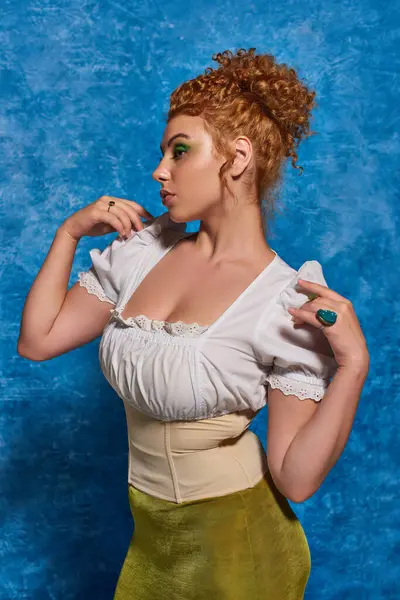 Joven pelirroja y curvilínea modelo en blusa blanca posando con las manos cerca de los hombros sobre fondo azul - foto de stock