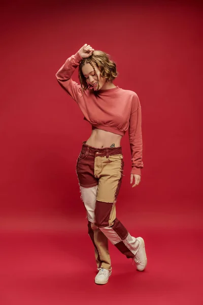 Glücklich stylische und junge Frau in Patchworkhosen und abgeschnittenem Langarm posiert auf rotem Hintergrund — Stockfoto