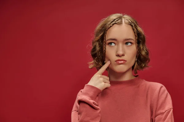Retrato de jovem mulher pensativa com cabelo ondulado curto tocando bochecha com o dedo no fundo vermelho — Fotografia de Stock