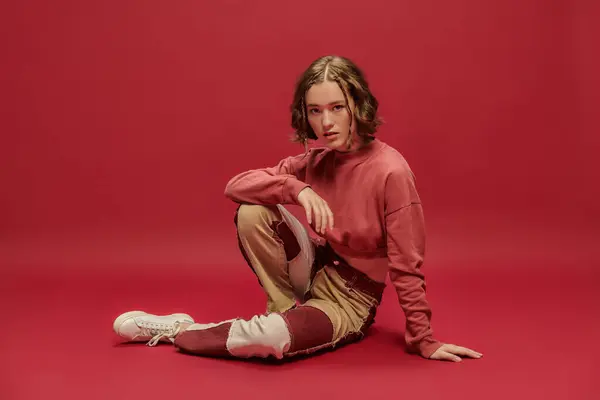Self-expression, young woman in patchwork pants and cropped long sleeve sitting on red backdrop — Stock Photo