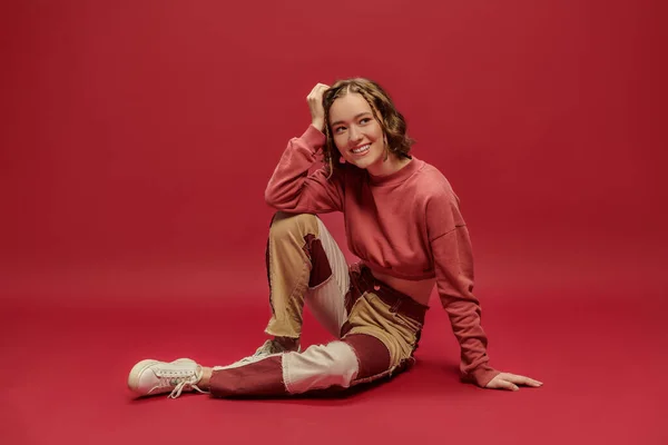 Expresión personal, mujer alegre en pantalones patchwork y manga larga recortada sentado sobre fondo rojo - foto de stock