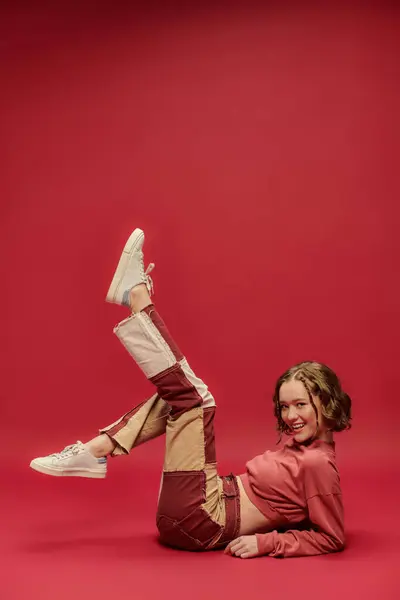Expresión personal, llena de alegría chica en pantalones patchwork y manga larga recortada sentado sobre fondo rojo - foto de stock