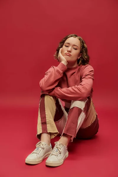 Bored and stylish gen z girl in patchwork pants and cropped long sleeve sitting on red backdrop — Stock Photo