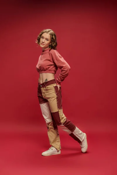 Estilo personal, chica bonita joven en pantalones patchwork y manga larga recortada posando sobre fondo rojo - foto de stock