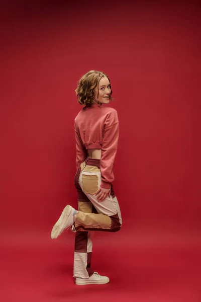 Estilo personal, joven chica alegre en pantalones patchwork y manga larga recortada posando sobre fondo rojo - foto de stock