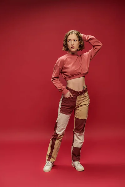 Jugendstil, junge hübsche Frau in Patchworkhosen und abgeschnittenem Langarm posiert auf rotem Hintergrund — Stockfoto