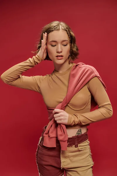 Young stylish woman in patchwork pants posing in tied jumper over long sleeve on red backdrop — Stock Photo