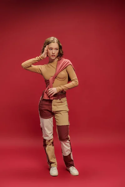 Young trendy woman in patchwork pants posing in tied jumper over long sleeve on red backdrop — Stock Photo