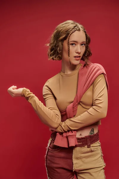 Trendige junge Frau in Patchworkhosen posiert im Pullover über abgeschnittenem Langarm vor rotem Hintergrund — Stockfoto