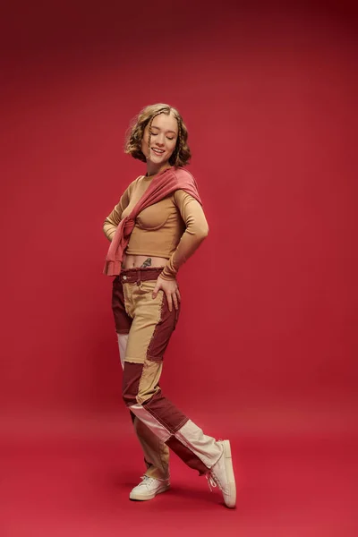 Positive jeune femme en patchwork pantalon posant en pull sur recadrée manches longues sur fond rouge — Photo de stock