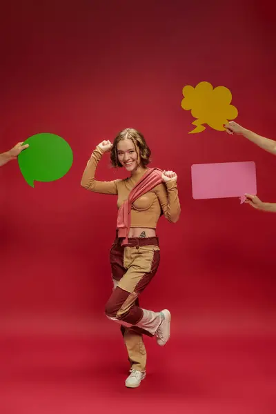 Femme joyeuse en patchwork pantalon et pull sur manches longues dansant près des gens avec des bulles de parole — Photo de stock