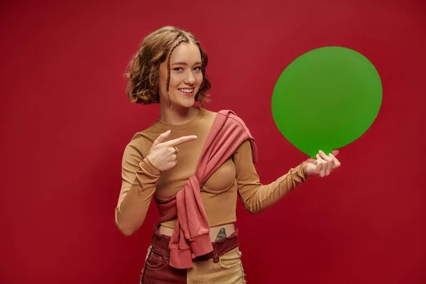 Mujer feliz en pantalones patchwork y puente sobre manga larga recortada apuntando a la burbuja del habla en rojo - foto de stock