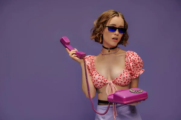 Молода жінка в модних сонцезахисних окулярах і обрізана зверху з серцями тримає ретро телефон на фіолетовому — стокове фото