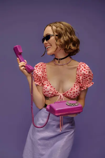 Happy young woman in trendy sunglasses and cropped top with hearts holding retro telephone on purple — Stock Photo