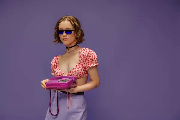 Mujer bonita en gafas de sol de moda y top recortado con corazones sosteniendo teléfono retro en púrpura - foto de stock