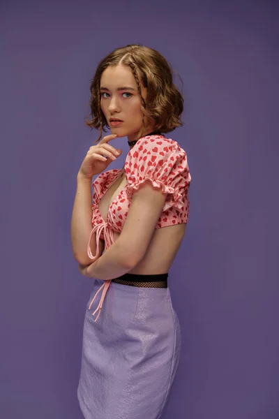 Nachdenkliche junge Frau in abgeschnittenem Top und Rock, die das Kinn auf lila Hintergrund berührt, persönlicher Stil — Stockfoto