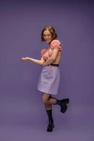 Souriant jeune femme en haut et jupe recadrée posant en bottes et collants résille sur fond violet — Photo de stock