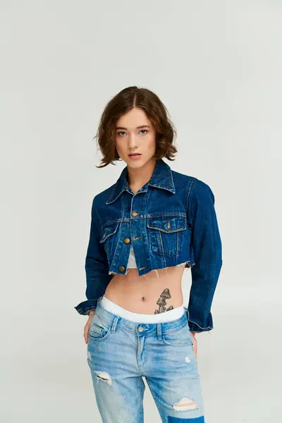 Stylische junge Frau mit Tätowierung posiert in abgeschnittener Jeansjacke und blauer Jeans auf grauem Hintergrund — Stockfoto