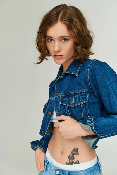 Stylisches Mädchen mit Tätowierung in abgeschnittener Jeansjacke posiert mit Hand in Jeanstasche auf grau — Stockfoto