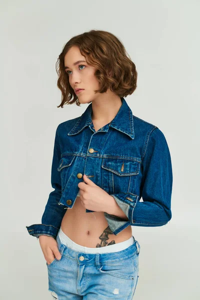 Модная девушка с татуировкой в обрезанной джинсовой куртке позирует с рукой в кармане джинсов на сером — стоковое фото