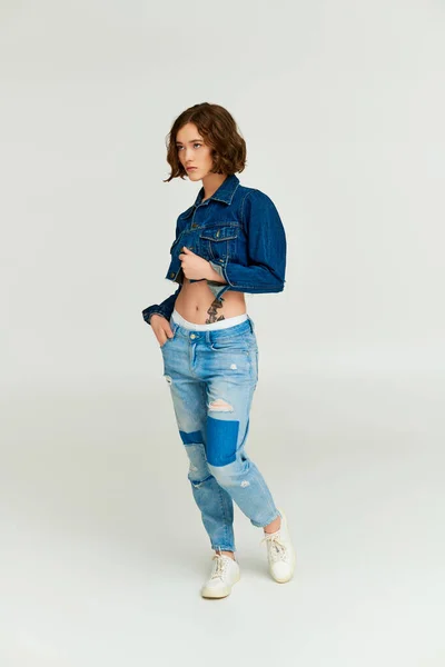 Красивая девушка в модных обрезанных джинсовой куртке позирует с рукой в кармане джинсов на сером фоне — стоковое фото