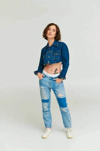 Chica de moda en chaqueta de mezclilla recortada posando con las manos en bolsillos de jeans sobre fondo gris - foto de stock