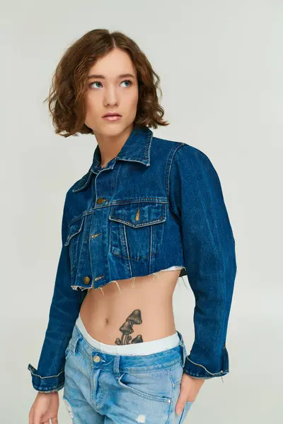 Attractive young woman with tattoo in cropped denim jacket and blue jeans posing on grey backdrop — Stock Photo