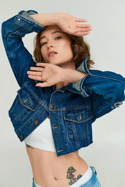 Attraente modello giovane con tatuaggio in giacca di denim ritagliato e jeans blu in posa su sfondo grigio — Foto stock
