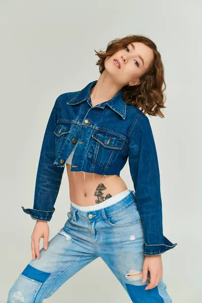 Auto-expresión, modelo de moda en chaqueta de mezclilla recortada y jeans azules posando sobre fondo gris - foto de stock