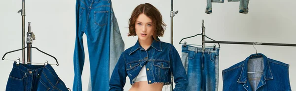 Hübsches Model in abgeschnittener blauer Jacke posiert inmitten von Jeanskleidung auf Kleiderbügeln vor grauem Hintergrund, Banner — Stockfoto