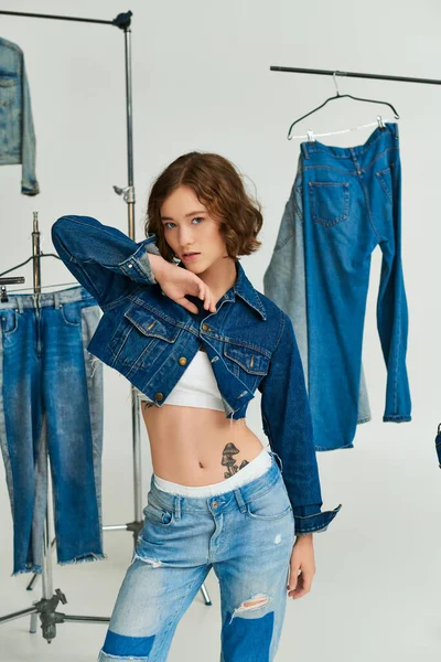 Bella giovane donna con tatuaggio posa in giacca blu ritagliato tra i vestiti di jeans su appendini su grigio — Foto stock