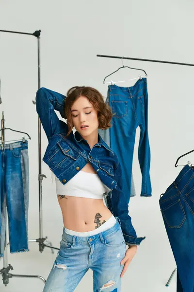 Charming young woman with tattoo posing in cropped blue jacket among denim clothes on grey backdrop — Stock Photo