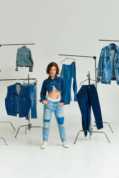 Fashion model in cropped jacket posing with hands in pockets among denim clothes on hangers on grey — Stock Photo