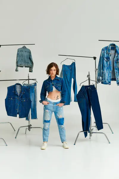 Modello moda in giacca ritagliata in posa con le mani in tasche tra i vestiti in denim su appendini grigi — Foto stock