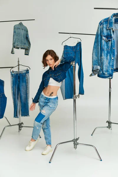 Stylish model with tattoo posing in cropped jacket and jeans among denim clothes on grey backdrop — Stock Photo