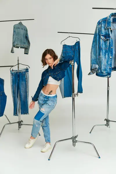 Modelo elegante con tatuaje posando en chaqueta recortada y vaqueros entre la ropa de mezclilla sobre fondo gris - foto de stock