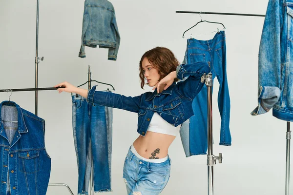Stylish model with tattoo posing in cropped jacket and jeans among denim clothes on grey background — Stock Photo