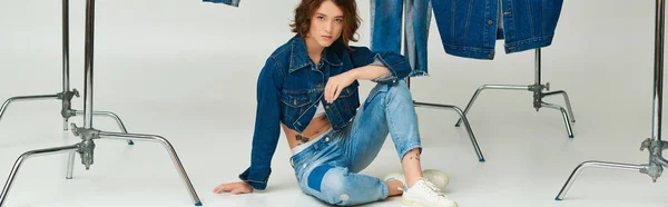 Stylish model with tattoo posing in cropped jacket and jeans sitting among denim clothes on grey — Stock Photo