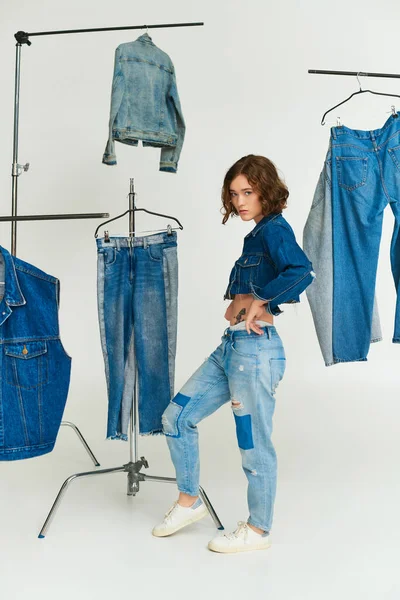 Stylisches junges Model mit Tätowierung in abgeschnittener Jacke und Jeans steht zwischen Jeanskleidung auf grau — Stockfoto